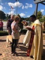 Priest in gold robe blessing a woman outdoors beside a dog under a sunny sky