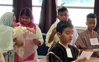 Children standing indoors and reading papers in a classroom, with a window and curtains behind them