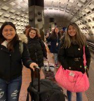 Four people with luggage walk through a tiled underground station, smiling at the camera.
