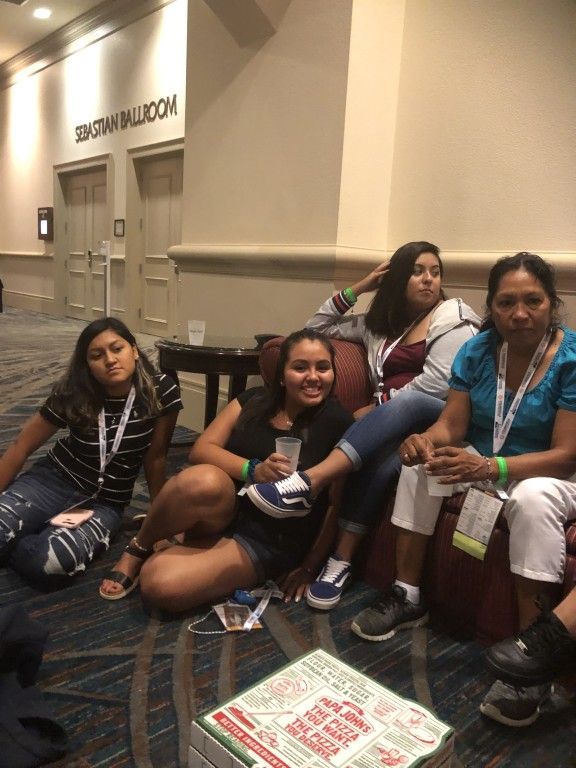 Four people sitting on a carpeted convention floor beside a wall and benches, one holding a sign.