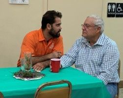 Two men talk at a table with a green tablecloth; one wears orange, the other a blue plaid shirt.