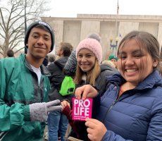 Three smiling people outdoors, holding a pink “PRO LIFE” sign in front of a building.