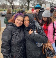 Two women smiling and hugging outdoors in winter coats, with a crowd behind them.
