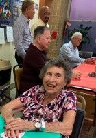 Smiling woman in floral top seated at a table in a community room with other people behind her