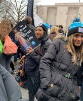 People in winter coats at a protest, one holding a sign, outdoors in cold weather