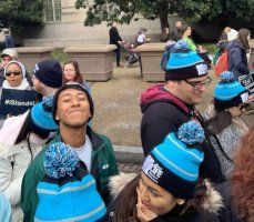 People wearing blue knit hats with pom-poms gathered outdoors in a crowd.