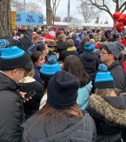 Crowd gathered outdoors in winter hats, facing a stage or speaker, with red balloons and a blue sign in the background
