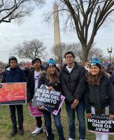 People holding protest signs in a park, with the Washington Monument in the background.
