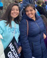 Two smiling friends standing outdoors, one in a light blue hoodie and one in a navy puffer vest.