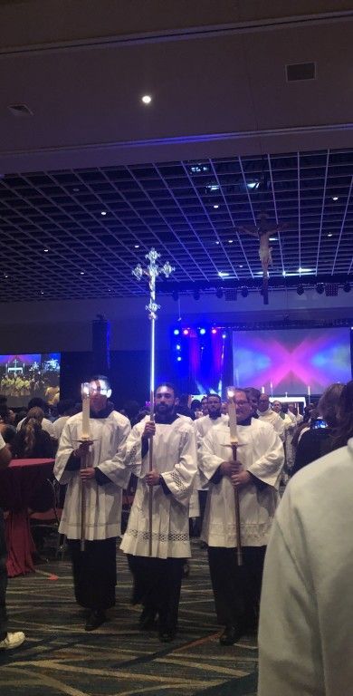 Four robed clergy stand in an indoor hall with purple stage lights and a crowd behind them.