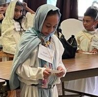 Child in a light green headscarf holding a small paper item at a table, with other children in the background