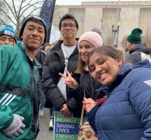 Group of four people posing outdoors in winter coats, smiling with a banner reading “WOMEN SAVING LIVES ENDING...”