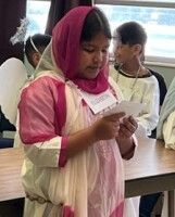 Child in pink and white clothes reading a note at a table, with another child in the background.