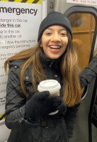 Smiling person in a black beanie and gloves holding a coffee cup on a subway train