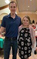 Two people posing indoors at a formal event, one in a navy shirt and jeans, the other in a black floral dress.