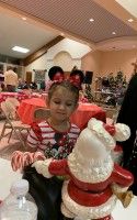 Child in red outfit sits at a table with a Santa decoration in a festive room.