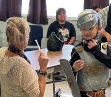 Child in knight costume holds a shield at a table while another person writes nearby in a room