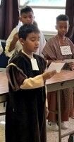 Children in traditional clothing reading from papers at a podium in a classroom
