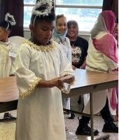 Child in a white ceremonial outfit stands at a classroom table while classmates watch nearby.