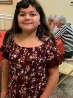 Smiling child in a burgundy floral top standing indoors with seated people in the background