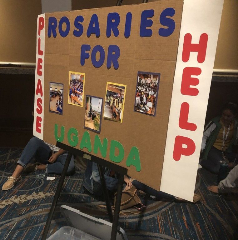 Handmade sign reading “Rosarie’s for Uganda” and “Please Help,” with small photos attached