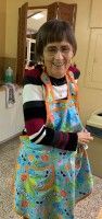 Smiling person in a colorful apron and striped shirt standing indoors with arms crossed.