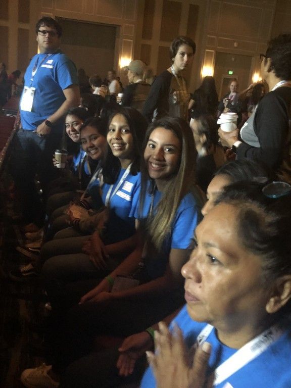 Audience seated at a conference, many wearing blue shirts and lanyards, listening attentively.