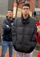 Two people standing outdoors by a brick building, one in a black jacket in front and one in a navy sweater behind.
