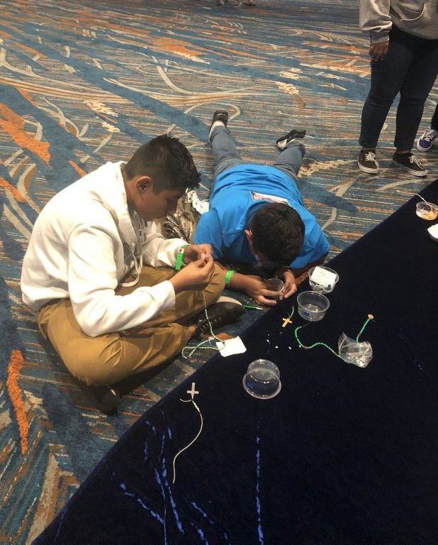 Two people crouch on a carpet, assembling a small device with wires and cups on a black table.
