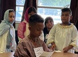Children standing around a table, watching a boy read from a sheet of paper indoors.
