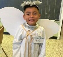 Child in a white angel costume with halo and wings, smiling indoors in front of a chalkboard.