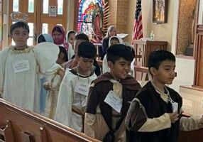 Children walking in a church aisle during a procession, wearing robes and name tags.