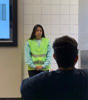 Presenter in a neon safety vest stands in front of a screen, addressing an audience in a classroom.