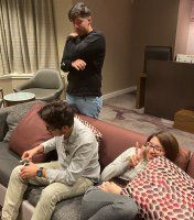 Three people in a living room, two sitting on a couch and one standing behind them, all looking at phones.
