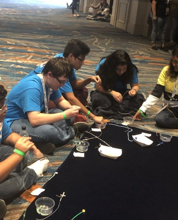 People sitting on the floor working with wires and small tools around a black tablecloth in a workshop setting