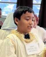 Person in a cream graduation gown and gold stole stands indoors, speaking with a microphone nearby.