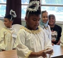 Children in white costumes with gold trim stand indoors during a performance, one holding a prop.