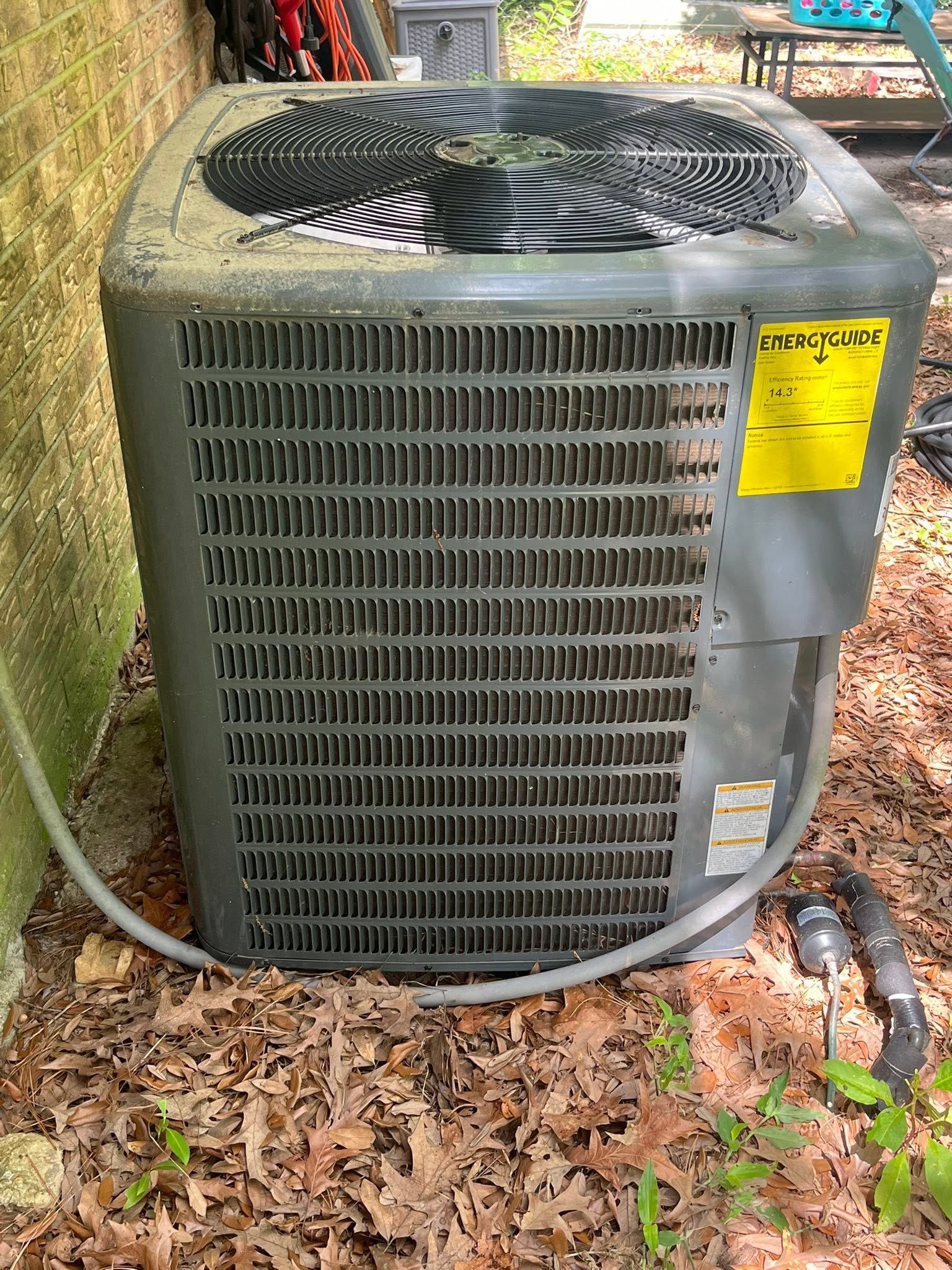 Outdoor air conditioner unit with a top fan, sitting beside a wall and covered in leaves.