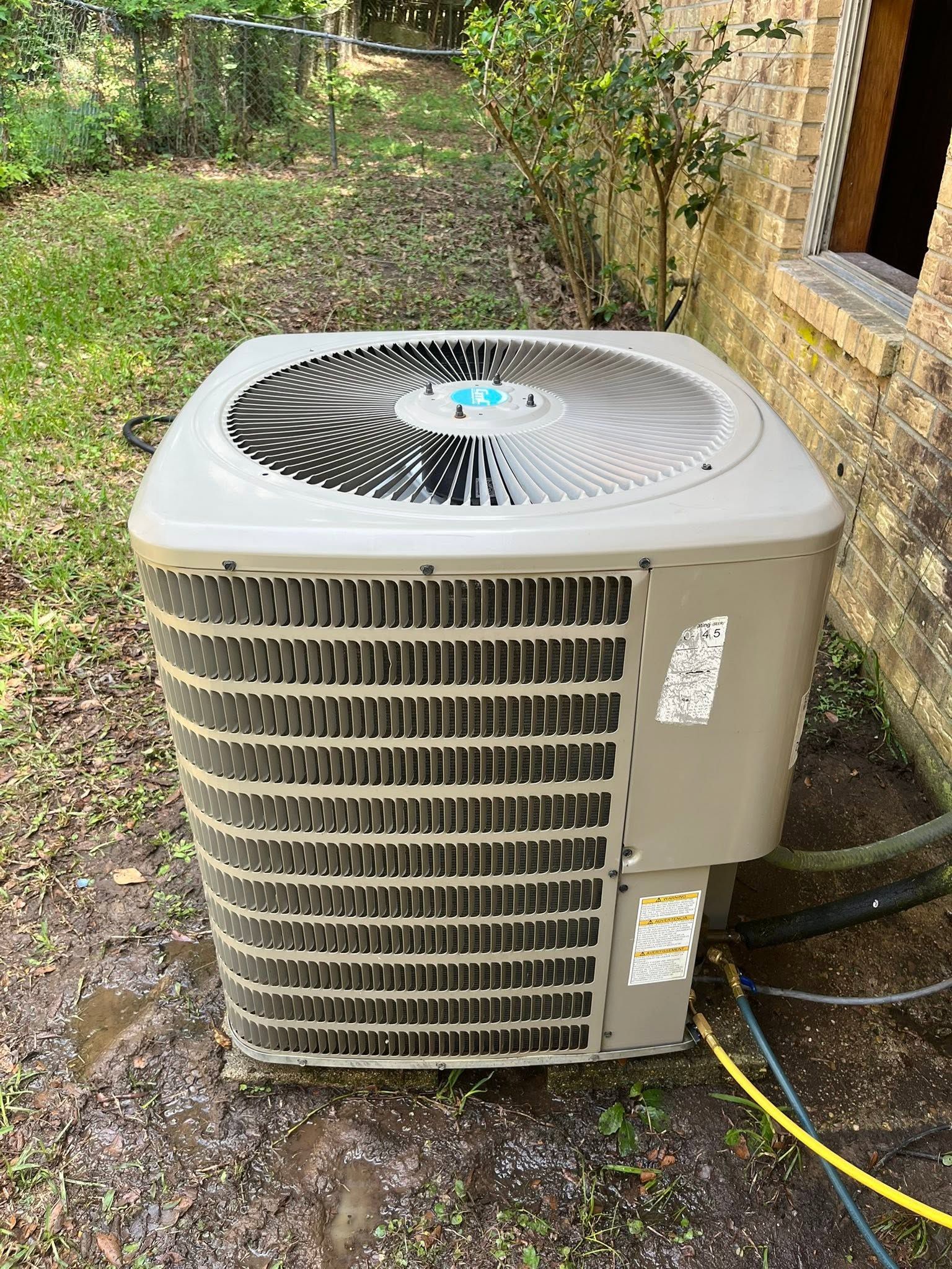 Outdoor HVAC unit beside a brick house, with hoses attached in a grassy yard.