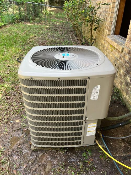 Outdoor air conditioner condenser unit beside a brick house, with hoses attached in a grassy yard