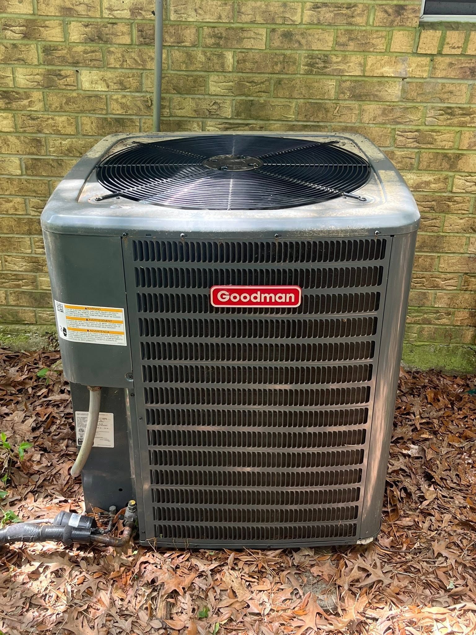 Outdoor Goodman air conditioner unit beside a brick wall, with leaves on the ground.