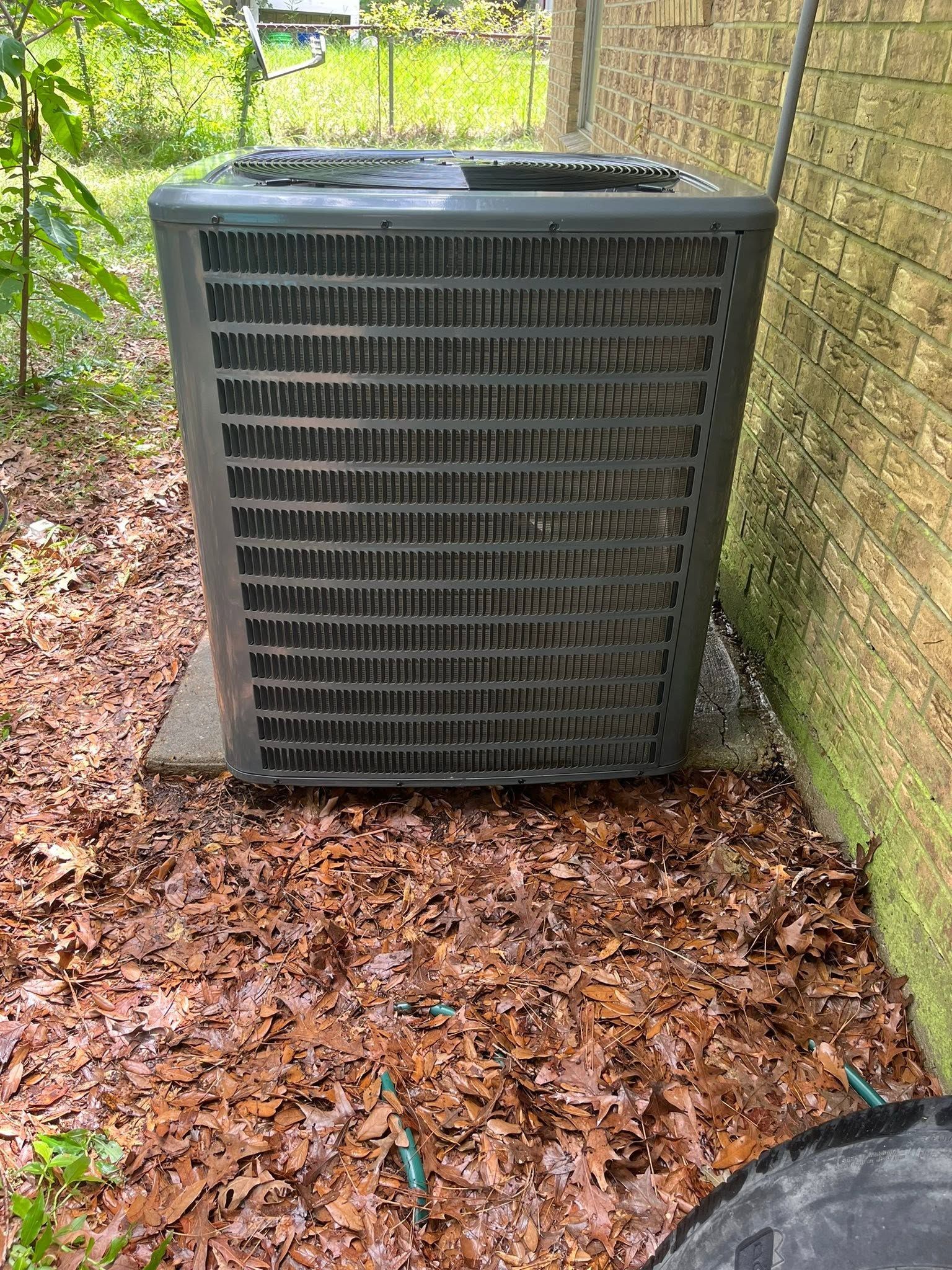 Outdoor HVAC unit beside a brick wall, surrounded by fallen leaves.