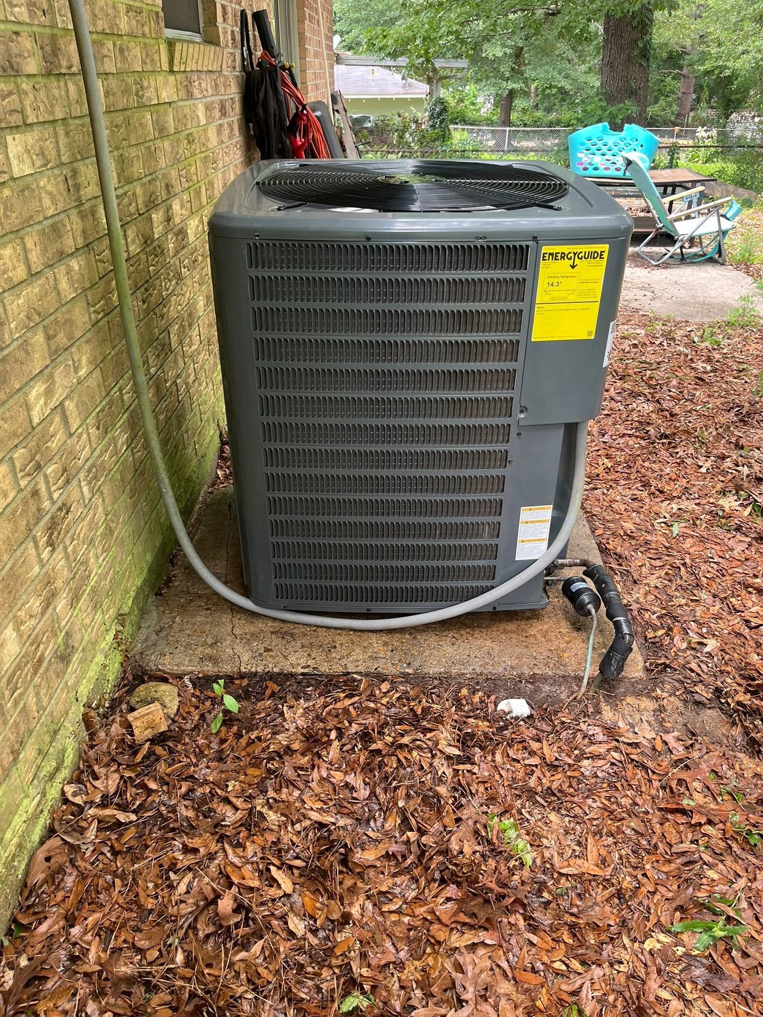 Outdoor air conditioning unit beside a brick wall, sitting on a concrete pad with leaves around it.
