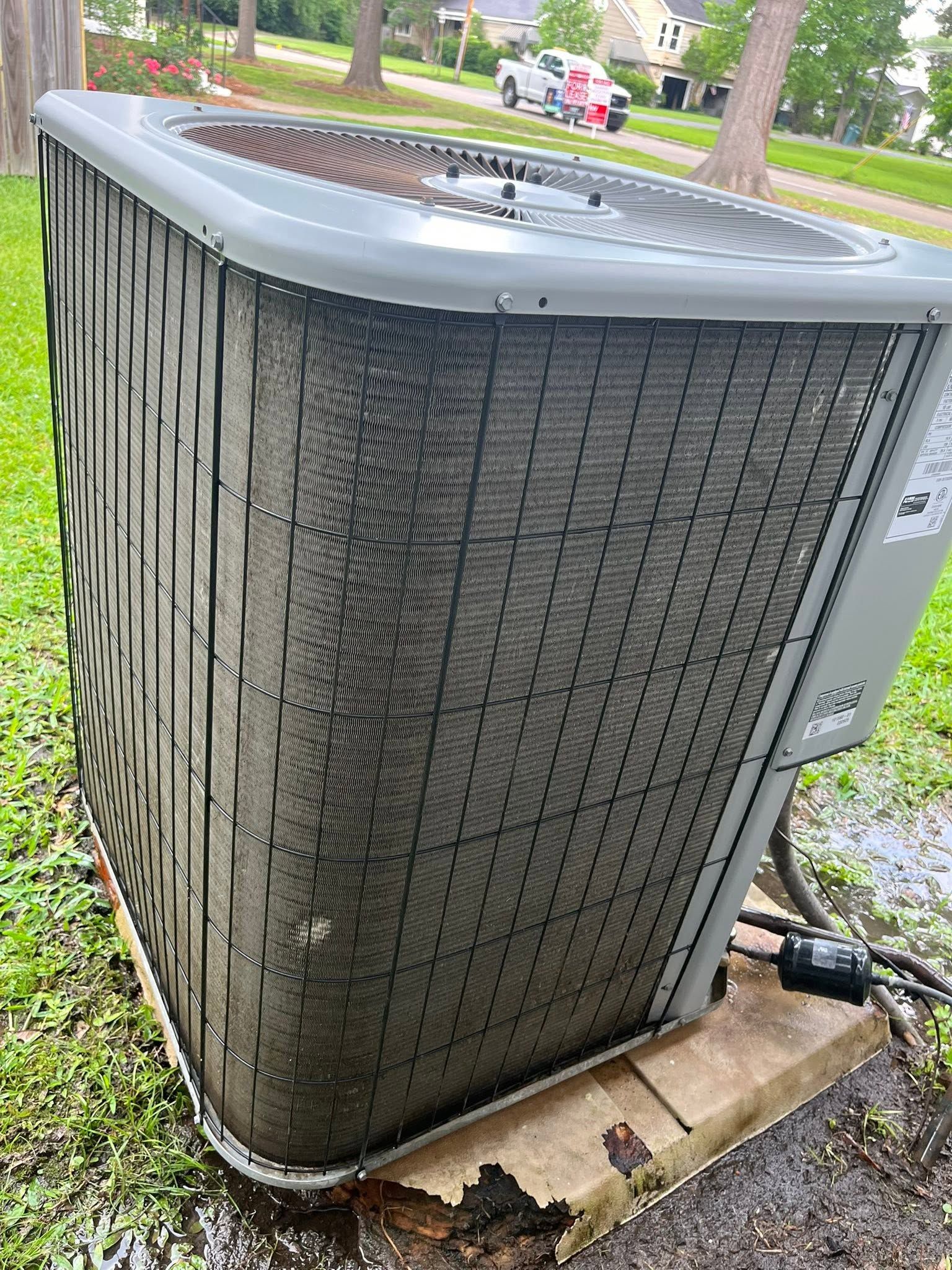 Outdoor HVAC condenser unit with dark, dirty coils beside a grassy driveway