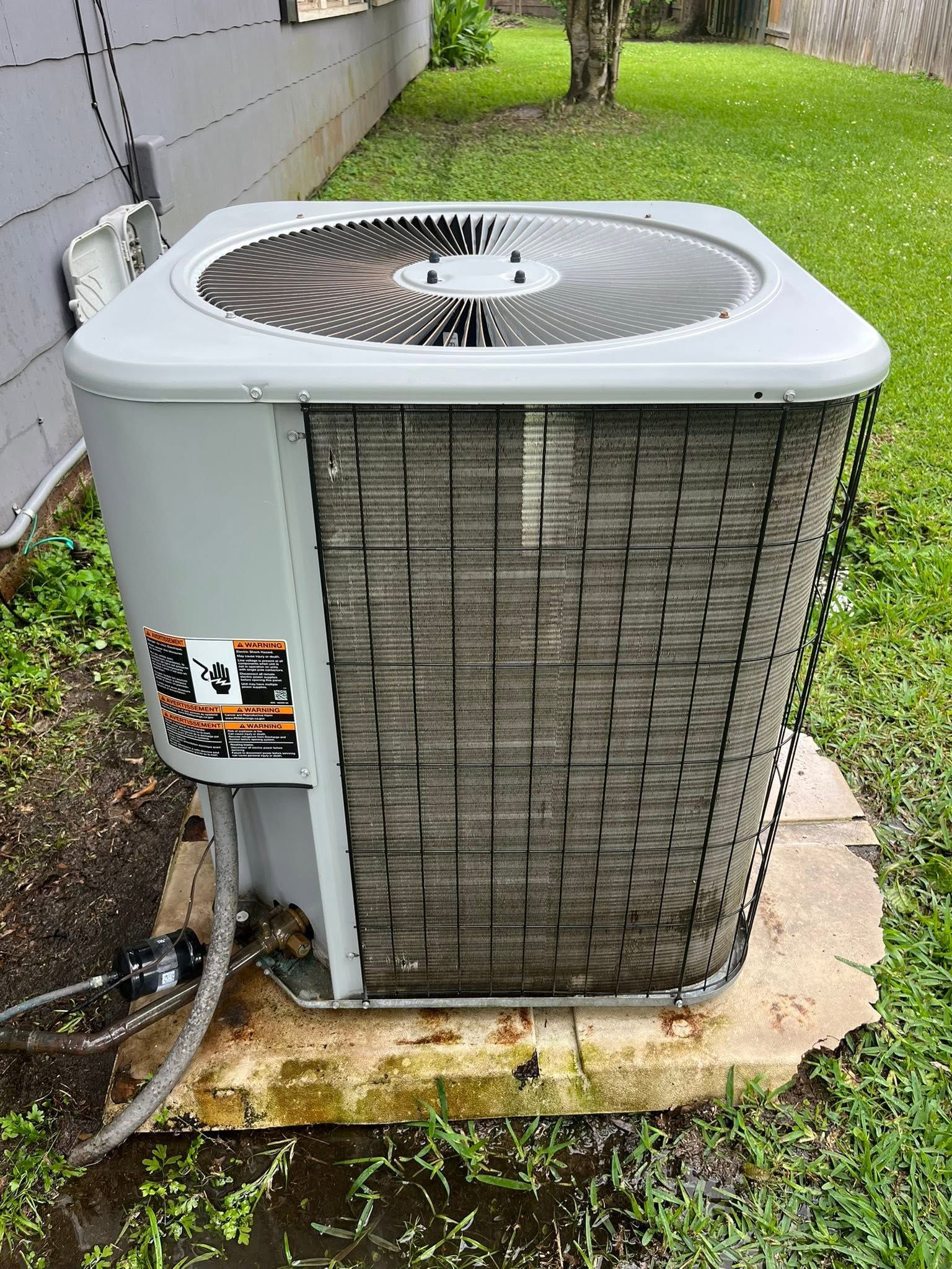 Outdoor air conditioner unit beside a house wall, with a large fan on top and a weathered metal grille.