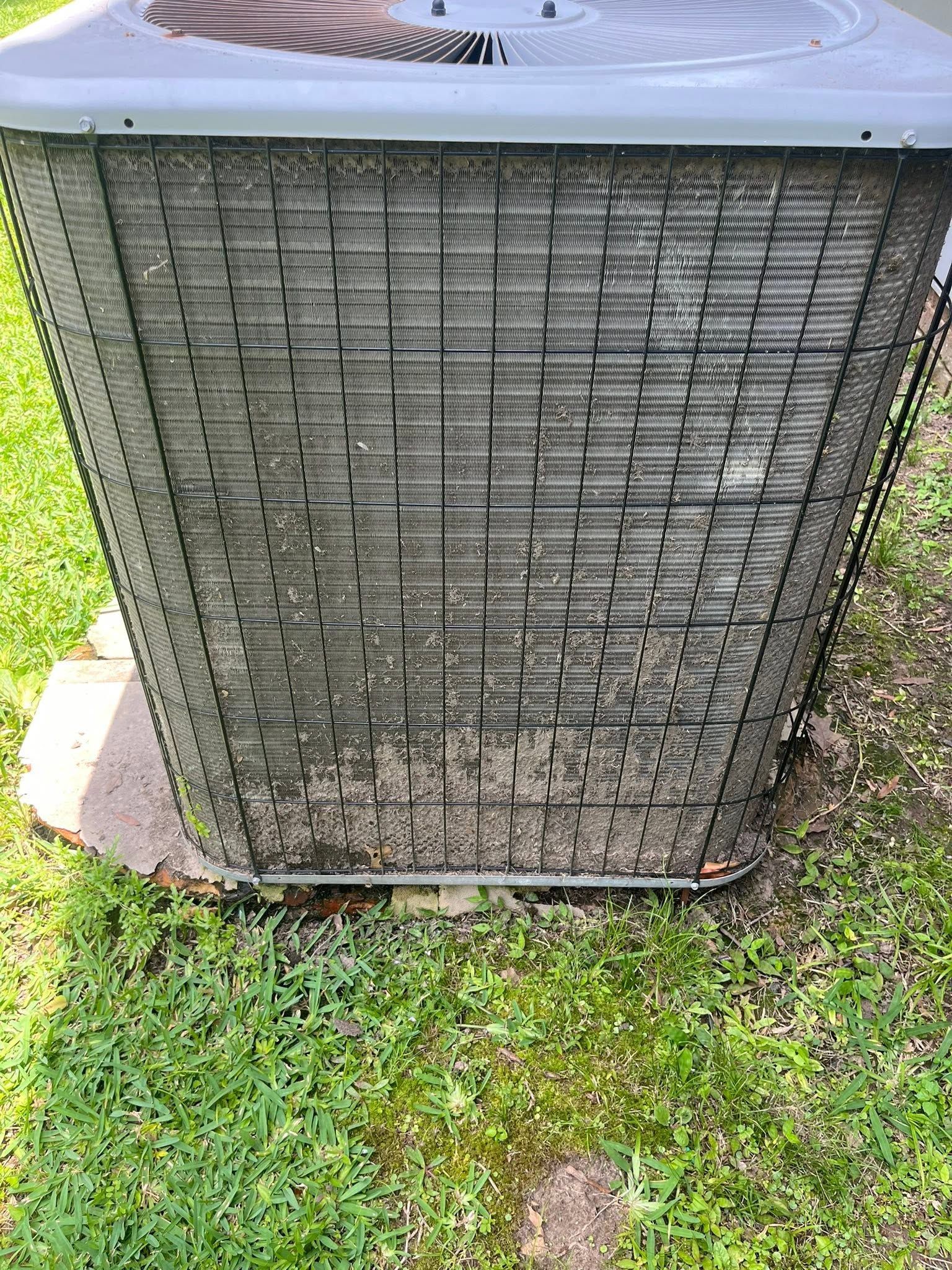 Outdoor HVAC condenser unit with a metal grille sitting on grass beside concrete and a house wall