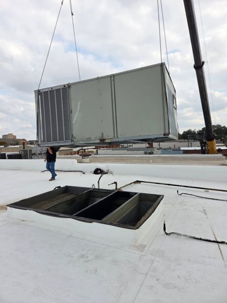 Crane lifting a large gray HVAC unit onto a white rooftop near an open skylight.