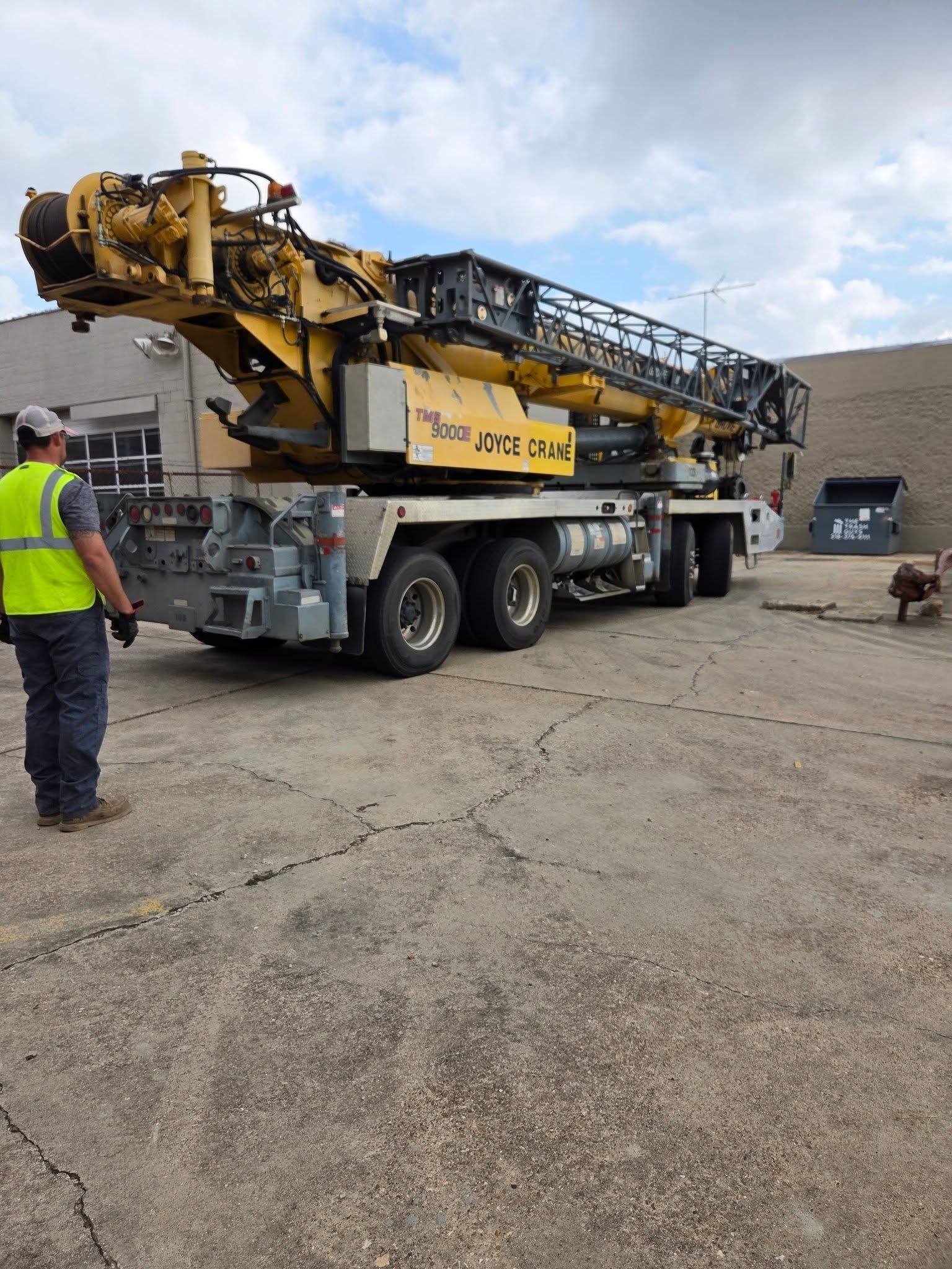 Yellow heavy industrial machine on a flatbed truck at a worksite, with a worker in a safety vest nearby