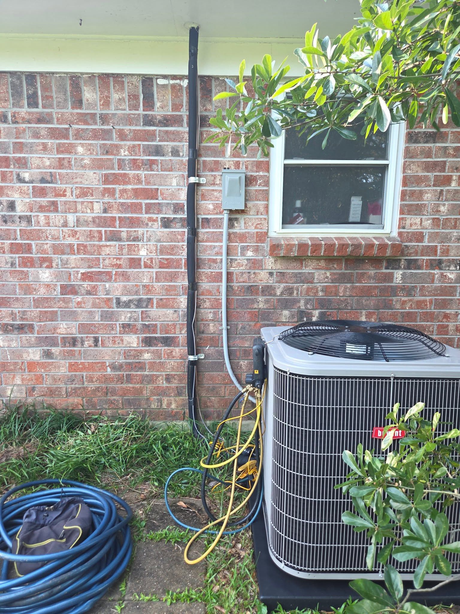 Outdoor AC unit beside a brick house wall, with hoses and an electrical box under a window.