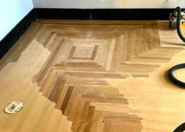 A wooden floor with a herringbone pattern is being sanded with a vacuum cleaner.